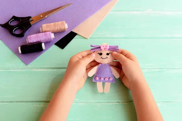 Ребенок держит в руках маленькую куклу. Ребенок показывает войлочную куклу. Ножницы, нитки и войлоки накрыты на деревянный стол. Простая художественная и ремесленная идея для детей — стоковое фото