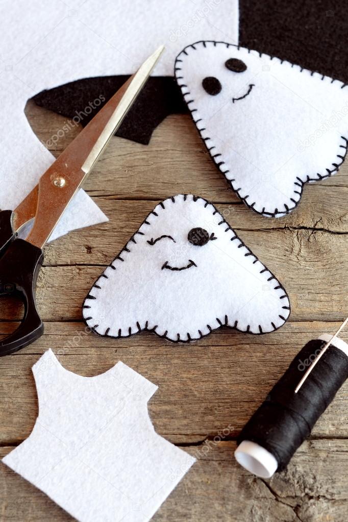 Cute white ghosts crafts, felt sheets, scissors, thread, needles on an old wooden background. Easy Halloween felt ghosts decor. Halloween sewing project for kids 