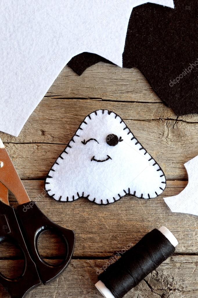 Cute felt Halloween ghost toy, scissors, black thread, scraps of felt on old wooden background. Supplies for ghost toy sewing. Halloween craft idea for kids. Closeup. Top view 