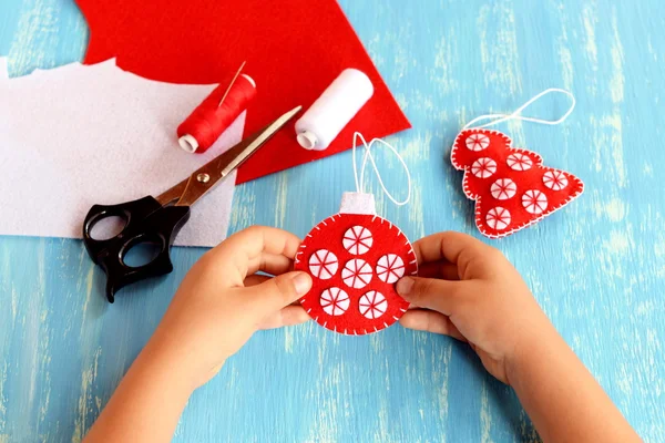 Küçük çocuk elinde bir Noel ağacı topu tutuyor. İşlemeli kırmızı ve beyaz top ve ağaç süslemeleri. Keçeli kumaş, iplik, iğne, makas mavi ahşap arka planda. Çocuklar Noel işçiliği. Anaokulu öğrencileri için Noel el dikiş atölyesi