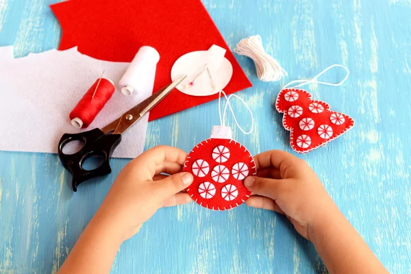 Elinde kırmızı bir Noel balosu süsü tutan bir çocuk. Çocuk kırmızı bir Noel balosu süslemesi gösteriyor. Noel ağacı deseni, makas, iplik, iğne, mavi arka planda keçe kırıntıları. Dikiş konsepti. Kış minimalizm sanatı
