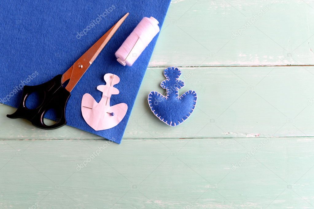 Home made anchor decor. Blue felt anchor is sewn with white thread, needle, paper pattern pinned on a flat piece of blue felt, scissors on a yellow background. Sewing lesson for children. Step 