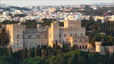 Rodos Yunanistan 'daki Büyük Üstat Sarayı' nın manzarası. Görkemli taş bina yeşil ağaçların arasında duruyor. Arka planda güneş ışığıyla yıkanmış bir şehir var..