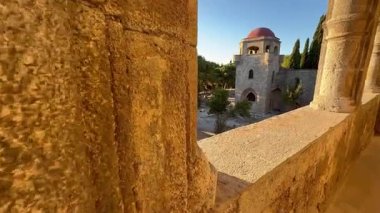 Rodos 'a git ve Yunanistan' daki Filerimos Manastırı 'nın taş mimarisini gözlemle. Kırmızı kubbeli bir kule kemerli taş geçitlerden görünen ağaçların arasında duruyor..
