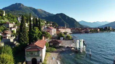 Selvi ağaçları ve berrak göl tarafından çerçevelenmiş sahil geçidi, çakıl taşları, villalar ve rıhtımlar gibi hafif dalgalar.