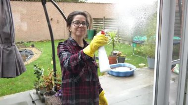 Eldiven giyen bir kadın cam veranda kapılarına temizlik solüsyonu sıkar. Arka bahçe ve açık teras camdan görülebiliyor. Temizlerken gülümsüyor..