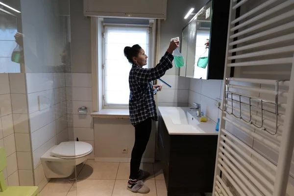 A woman sprays cleaning solution onto a mirror and prepares to wipe it with a squeegee. She is cleaning the bathroom of a home.