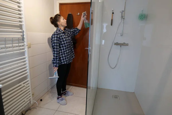 A woman sprays cleaning solution onto the glass door of a shower in a bathroom. She holds a blue squeegee in her other hand and wears slippers.