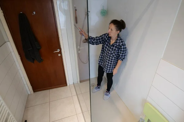 A woman in a blue plaid shirt and black leggings cleans inside a glass shower stall with a cleaning tool. She stands in a bathroom with light colored tiles.