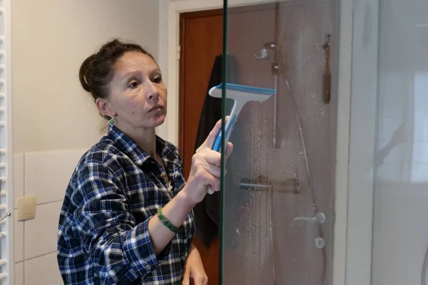 A middle-aged woman wearing a plaid shirt is cleaning a glass shower door in her bathroom. She is using a squeegee to remove water and soap scum.