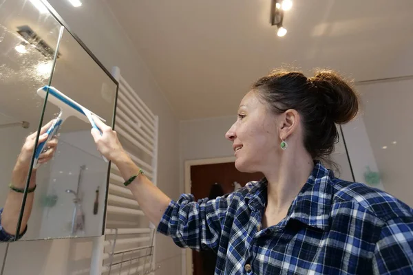 A woman cleans a mirror in the bathroom. She is using a blue squeegee to wipe the mirror. The mirror is large and the bathroom is well lit. She is smiling during the chore.