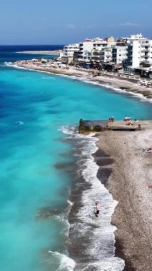 Genel bakış açıları kıyı şeridi binaları ve Ixia Beach Rodos boyunca parlak turkuaz suyu gösteriyor. İnsanlar sahilde dinlenir ve güneşli havanın tadını çıkarırlar..