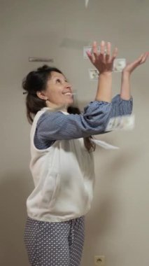 Orta yaşlı, desenli pantolonlu, mavi gömlekli ve beyaz yelekli bir kadın, Amerika Birleşik Devletleri para birimini heyecanlı bir şekilde kapalı havada yakalıyor. Gülümsüyor ve mutlu görünüyor..