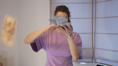 Mor gömlekli genç bir kadın evde dolar banknotları saçıyor. Gülümsüyor ve servetinden zevk alıyor gibi görünüyor ya da ev ortamında zenginmiş gibi davranıyor..