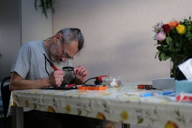 Olgun bir adam yemek masasında lehimli gözlük takıyor. Tahtayı incelemek için bir büyüteç kullanıyor. Masanın üzerinde çeşitli araç ve parçalar var.
