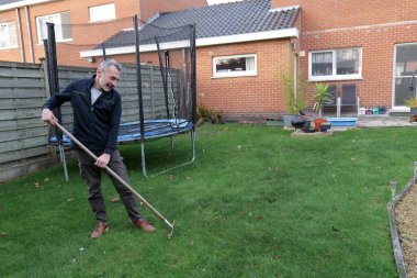 Bir adam yeşil çimlerin üzerinde duruyor ve yaprakları yapraklarla tırmıklıyor. Gündüz vakti kırmızı tuğladan bir ev ve trambolini olan bir evin arka bahçesinde..