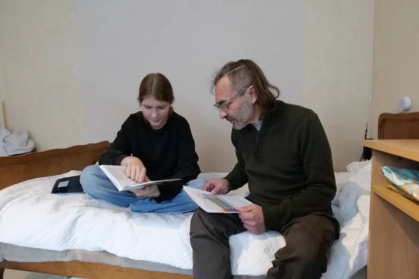 Bir baba ve kızı bir yatakta oturmuş okul malzemelerini gözden geçiriyorlar. Kız bir kitaba bakıyor ve adam renkli ve desenli kağıtları inceliyor..