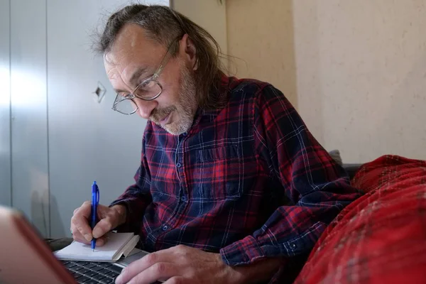 Yetişkin bir erkek gözlüklü ve kırmızı fanila gömlekli evde çalışıyor. Ayrıca küçük bir not defterine mavi kalemle not alırken bir dizüstü bilgisayar kullanıyor. Kanepede oturuyor..