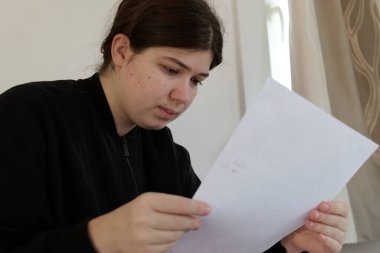 Siyah ceketli genç bir kız pencerenin yanındaki parlak bir odada elinde tuttuğu belgeyi okurken aşağıya bakıyor..