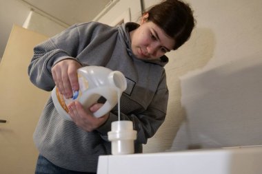 Genç bir kadın, çamaşır odasındaki çamaşır makinesine plastik bir konteynırdan sıvı deterjan döküyor..