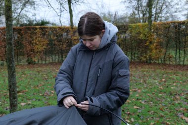 A teenager in a grey jacket is assembling a tent outside on a cloudy day. She is connecting the tent poles to the tent fabric.