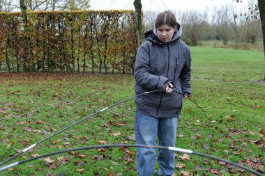Gri ceketli genç bir kadın dışarıda çadır direklerini birbirine bağlıyor. Mekan çimlerin üzerinde yapraklar olan bir tarlaya benziyor..