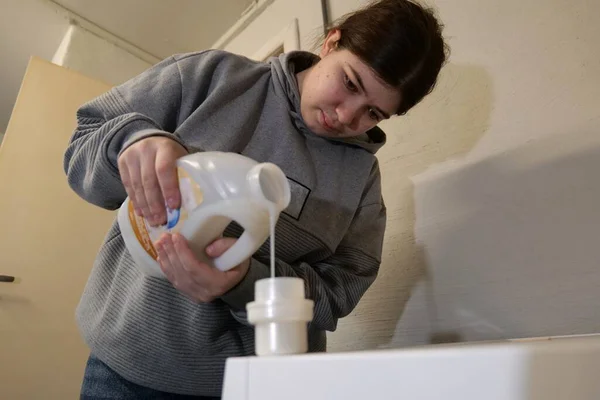 Genç bir kadın, çamaşır odasındaki çamaşır makinesine plastik bir konteynırdan sıvı deterjan döküyor..
