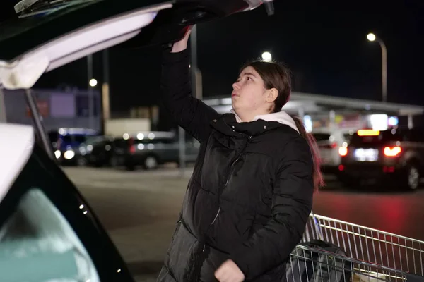 Siyah ceketli bir kadın alışveriş arabasından alışveriş çantalarını geceleri otoparktaki bir arabanın bagajına yüklüyor..