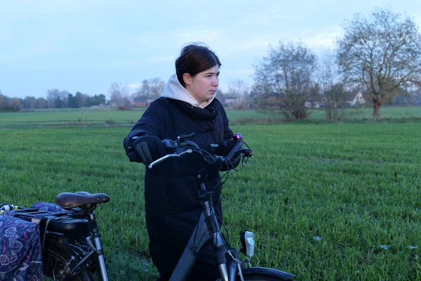 Paltolu genç bir kadın yeşil bir alanda iki bisikletle ayakta duruyor. Gökyüzü gri ya sabahın erken saatleri ya da alacakaranlığın..