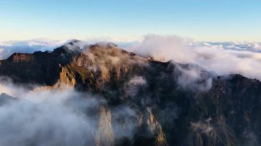 Büyüleyici Madeira dağ zirveleri güzel bir gündoğumu sırasında bulutlardan oluşan bir battaniyenin üzerinde yükselir. Altın ışık, şafak vakti engebeli manzarayı aydınlatır..