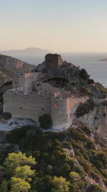 Altın saat ışığı, antik Kritinia Kalesi 'ni Rodos Yunanistan' daki bir uçurumun üzerinde yıkıyor. Ege Denizi ve engebeli araziler uzanıyor.