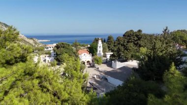 Beyaz bir manastır ve Rodos Yunanistan 'daki köy boyunca panoramik manzara. Yeşil ağaçlar, açık mavi Ege Denizi ve güneşli gökyüzüne karşı yapılar oluşturuyor..