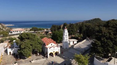 Rodos Yunanistan 'daki Tsambika Manastırı' nın genel manzarası. Beyaz binalar, kırmızı çatılar çan kulesi ve yemyeşil ağaçlar durgun mavi Ege Denizi 'nin yanında duruyor..