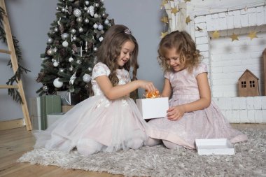 İki kız şöminenin yanında çelenk kutusunu tutuyor. Noel ve yeni yıl için dekore edilmiş kart. Yeni yıl arkaplanında yerde oturan güzel elbiseli kız kardeşler.