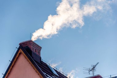 Soğuk kış havasında bacalı çatı açık mavi gökyüzüne duman yayıyor. Sis Çekimi. Hava Kalite Kısıtlamaları. Yakma Yasağı