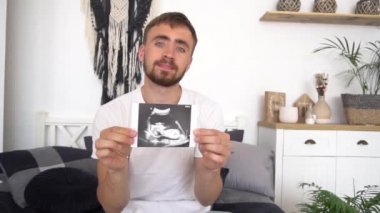 Ağır çekim hamile adam evdeki koltukta oturuyor ve ultrason görüntüsü gösteriyor.