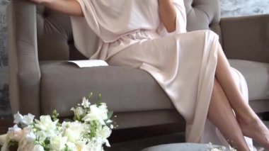 İpek kemerli beyaz penyuar giyen bir kadın kanepeye oturur.