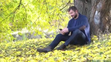 Kırk yaşında bir adam sonbaharda dışarıda otururken cep telefonuna bakıyor.