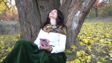 Telefonu olan bir kadın, sarı yaprakların arasında bir ağacın altında oturuyor. Sonbahar havası.