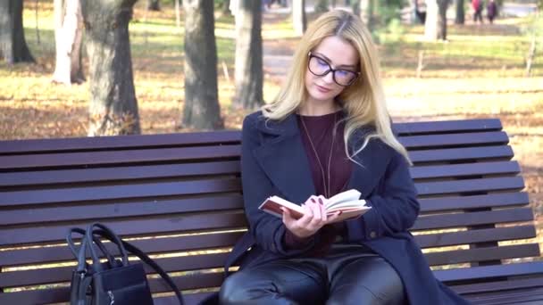 4K迷人的时尚女孩坐在秋天公园的长椅上看书 — 图库视频影像