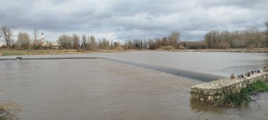 İspanya, Burgos 'ta bulutlu bir kış gökyüzünün altında küçük bir taş barajı olan hızlı akan bir nehrin manzarası.