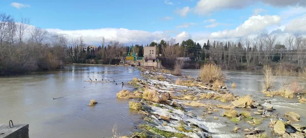 Logroo, İspanya 'da bulutlu bir gökyüzünün altındaki eski bir binanın önündeki taştan bir baraj ve küçük şelaleli nehir manzarası.