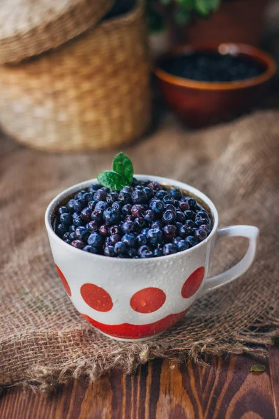 多汁蓝莓在老式杯前面查看静物。长满鲜花和篮子乡村背景. 免版税图库图片