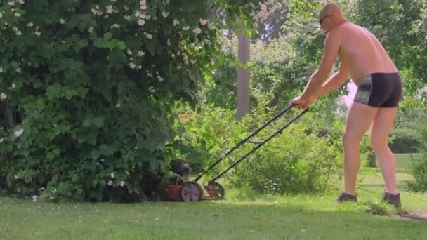 Взрослый мужчина в купальных штанах стрижет траву на лужайке в Эстонии. — стоковое видео