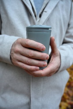 Bir adamın ellerinin basit bir seyahat kupasını tutarken estetik, minimalist görüntüsü nötr tonları ve modern sonbahar kıyafetlerini vurguluyor..