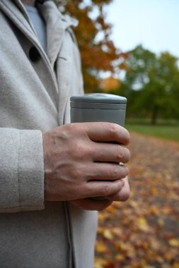 Tarafsız giyinmiş bir adam sıcak bir kupa tutuyor, sonbahar parkında bir yürüyüş sırasında durgunluk ve sakinlik anını yakalıyor. Ruh sağlığına odaklan.