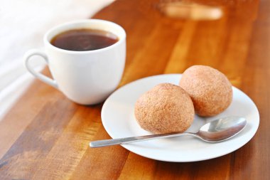 Beyaz bir fincan sade kahve ve ahşap tepside iki yuvarlak hamur işi. Sıcak kahvaltı ya da hafif doğal ışıklandırmalı atıştırmalık konsepti.