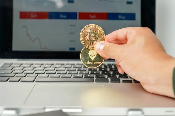 Man holding two physical currencies: Bitcoin and Ethereum in front of laptop with a background of stock market charts. Concept of new investments in virtual currencies 