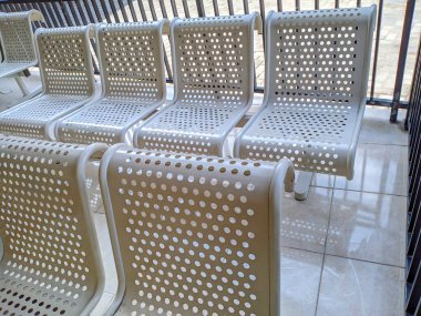Boş, delikli, açık renkli metal bir sıra bekleme koltuğu ya da fayanslı bir yerde, muhtemelen kapalı bir bekleme salonunda..