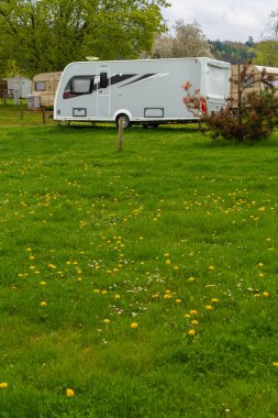 Çek Cumhuriyeti 'nde sakin bir bahar kampında beyaz, modern bir karavan. Yeşiller yemyeşil çimenler, çiçek açan karahindibalar ve ağaçlar dışarıda huzurlu bir yolculuk ortamı yaratır. Görünürde kimse yok, boşluğu kopyala.